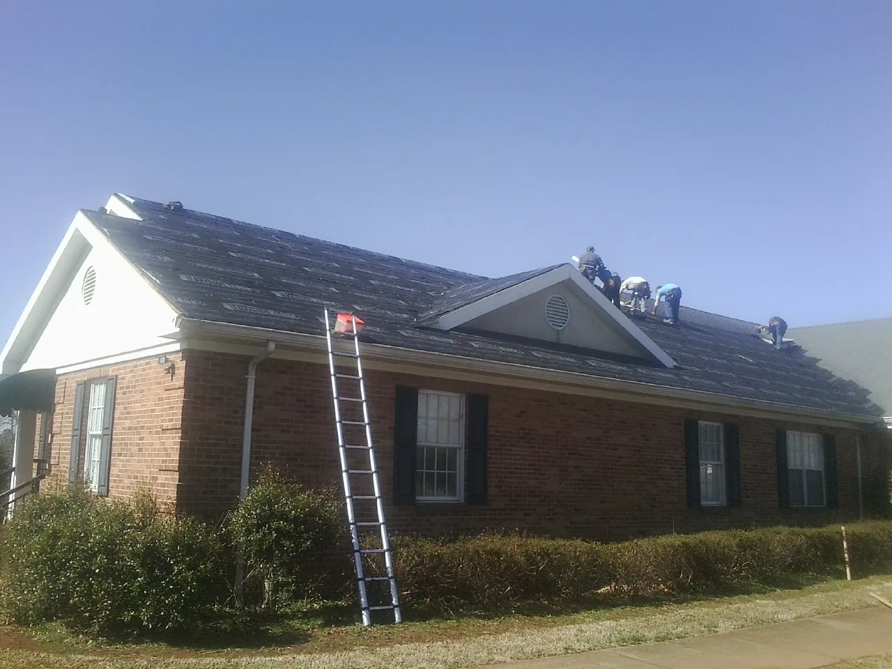 Crew tearing shingles off a brick ranch roof with a ladder set — Emergency Roof Repair work by our crew in Imlay