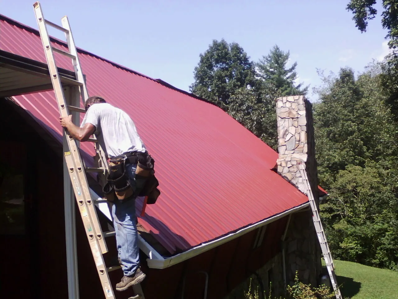 Roofer on a ladder beside a red metal roof and stone chimney — Soffit & Fascia Repair by Roofing Brothers in Imlay